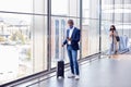 Businessman With Suitcase Standing By Window On Concourse At Railway Station With Mobile Phone