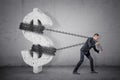 A businessman tugs at a chain trying to move a large concrete dollar sign from its place.