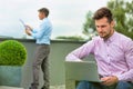 Young business man using his laptop while the other man reads newspaper in the background