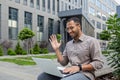 Businessman using laptop for video call, man sitting on bench outside office building, waving to interlocutor, greeting Royalty Free Stock Photo