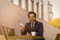 Businessman using mobile phone outdoors. Successful man toothy smiles holding smartphone while sitting at table with cup Royalty Free Stock Photo