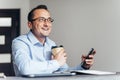 Businessman using phone, holding cup of coffee, sitting at his desk in modern office Royalty Free Stock Photo