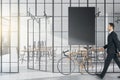Businessman walking in modern glass partition and bike in stylish coworking office interior with empty black mock up poster and