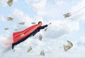 A businessman wearing a red superhero cape flying through the clouds following a dollar bill.