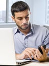 Businessman working on computer laptop using mobile phone at office desk in front of skyscraper window