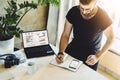 Businessman writes with pen in notebook while looking at smartphone. On table laptop with graphs, charts, diagrams on screen. Royalty Free Stock Photo