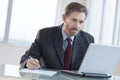 Businessman Writing Notes While Using Laptop At Desk