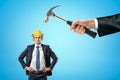 Businessman in yellow hard hat with ear defenders, standing with hands on hips, and big hand holding hammer to his head. Royalty Free Stock Photo