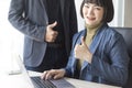 Businesswoman and businessman showing thumbs up sign with computer on wooden table in office background. Royalty Free Stock Photo