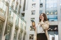 Businesswoman drinking hot coffee on the go in the middle of the street standing on background of office center talking on her Royalty Free Stock Photo