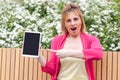 Businesswoman in elegance style sitting in bench on park, holding tablet empty screen and pointing finger to device with opened