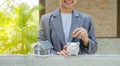 Businesswoman hands putting coin into piggybank, the home model stands in front of home background, Saving money for buy real