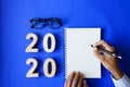 Businesswoman handwriting Happy New year 2020 Planning Goals on notebook with wooden number and glasses on classic blue paper