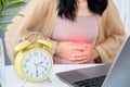 Business woman having stomach pain sitting at office with alarm clock on desk late for lunch time Royalty Free Stock Photo