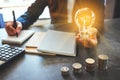 Businesswoman holding a lightbulb while taking note on notebook with coins stack on table