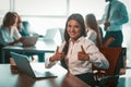 Businesswoman shows thumbs up in office. Alright gesture from young smiling girl. Group of businesspeople work on