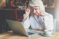 Businesswoman sits at table in front of laptop and looks closely at monitor, raising her glasses. Pensioner freelancer Royalty Free Stock Photo