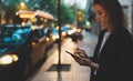 Businesswoman stands by busy road of evening city and calls smartphone of internet technologies, elegant girl waiting for taxi