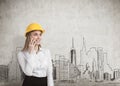 Businesswoman wearing a yellow hard hat and talking on her smartphone while standing near a concrete wall with a city panorama. Royalty Free Stock Photo