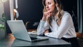 Businesswoman in white shirt is sitting in office at table in front of computer and pensively looks at screen of laptop Royalty Free Stock Photo