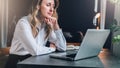 Businesswoman in white shirt is sitting in office at table in front of computer and pensively looks at screen of laptop Royalty Free Stock Photo