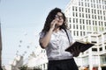 Businnes woman in office style clothing talking on the mobile phone with client and holding in hand her notebook