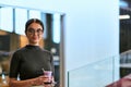 In a bustling modern office a businesswoman in glasses juggles her tasks, sipping coffee and using her smartphone