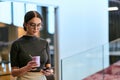 In a bustling modern office a businesswoman in glasses juggles her tasks, sipping coffee and using her smartphone