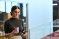 In a bustling modern office a businesswoman in glasses juggles her tasks, sipping coffee and using her smartphone