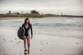 Busty female surfer strolling along wet sand.