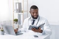Busy african doctor working with laptop onlinr, checking his notebook Royalty Free Stock Photo