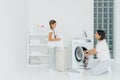 Busy brunette woman loads washing machine with dirty clothes, her little daughter helps, stands near basket and sorts laundry.