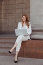 Busy business woman in white suit with laptop sitting outdoors at classical pillar. Investing, working remotely