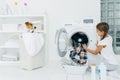 Busy child does laundry work, empties washing machine, cleaned clothes in basin uses detergents, little pedigree dog in basket. Royalty Free Stock Photo