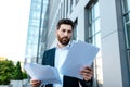 Busy confident young businessman with beard in suit works with documents near office building