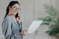 Busy female entrepreneur has telephone conversation, busy with accounting work, looks through paper documents, checks information Royalty Free Stock Photo