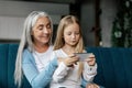 Busy glad caucasian little girl and elderly granny watch video, enjoy online game on smartphone, use new app