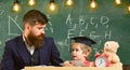 Busy kid studying at school. Teacher in formal wear and pupil in mortarboard in classroom, chalkboard on background Royalty Free Stock Photo