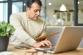 Busy latin middle aged businessman holding document envelope while working using laptop at home