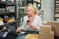 Busy mature woman, small business owner smiling and drinking coffee while working on the laptop, sitting at the desk in