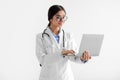 Busy millennial indian woman doctor in white coat and glasses working on laptop, isolated on white background Royalty Free Stock Photo