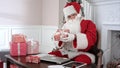 Busy Santa Claus preparing presents using laptop and digital tablet, sorting his letters and receiving a gift from a