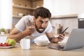 Busy serious adult caucasian man with beard in t-shirt eats porridge, uses smartphone, watches video on laptop