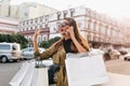 Busy shopaholic girl with bags talking on phone and waiting bus in city. Outdoor portrait of adorable young woman in Royalty Free Stock Photo