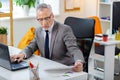 Busy tired grey-haired man in clear glasses reading papers and checking laptop