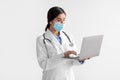 Busy young indian lady doctor in white coat and protective mask working on laptop isolated on light background Royalty Free Stock Photo