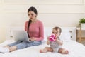 Busy young mother working on laptop while her little kid drinking water from bottle, sitting on bed, focus on child Royalty Free Stock Photo