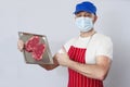 Butcher in blue face mask and red apron holding metal tray with two fresh premium strip loin steaks. Grey background. Meat