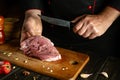 A butcher cuts veal with a knife in the kitchen for grilling or barbecue. Peppers and spices on the kitchen table to prepare a Royalty Free Stock Photo