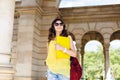 Buttom view pretty brunette girl in sunglasses walking with yellow laptop on archway background. She wears vinous bag
